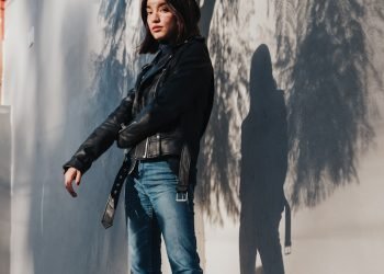 woman in black leather jacket and blue denim jeans standing beside white wall