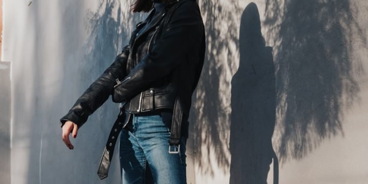 woman in black leather jacket and blue denim jeans standing beside white wall