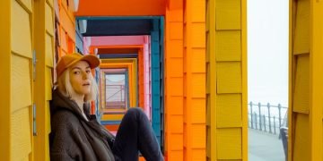 woman in brown jacket and blue denim jeans sitting on yellow wooden door during daytime